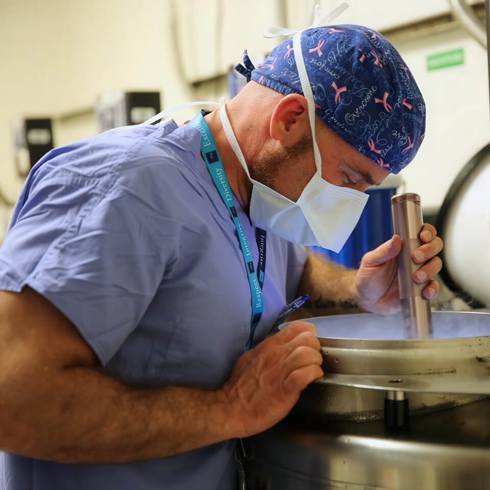 Embryologist in surgical scrubs and mask handling a cryopreservation tank with liquid nitrogen in a laboratory.