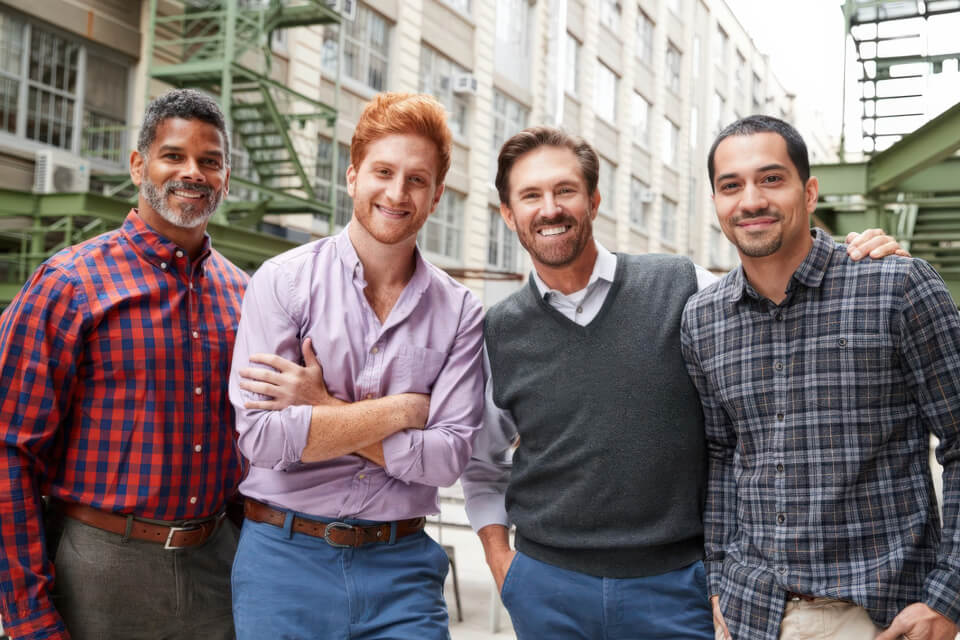 Four adult men standing together outdoors smiling at the camera.