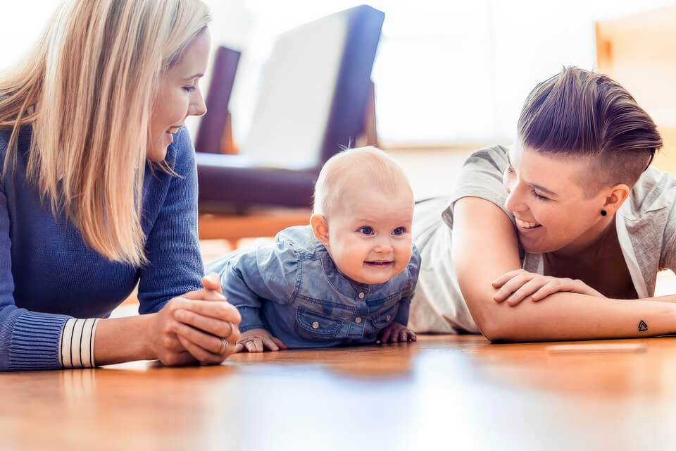 Two mothers lying on the floor smiling at their baby between them in a home setting.
